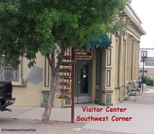 Tombstone Visitor Center Entry, tree on corner & directional arrow signs for town attractions. Bench around the corner by the Tombstone Art Gallery.